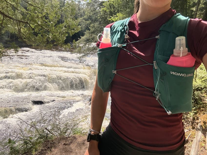 Trail runner with Salomon vest and water bottles standing in front of a waterfall. 